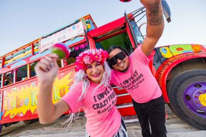 Two cheerful people in pink shirts dance on a beach near a colorful bus.