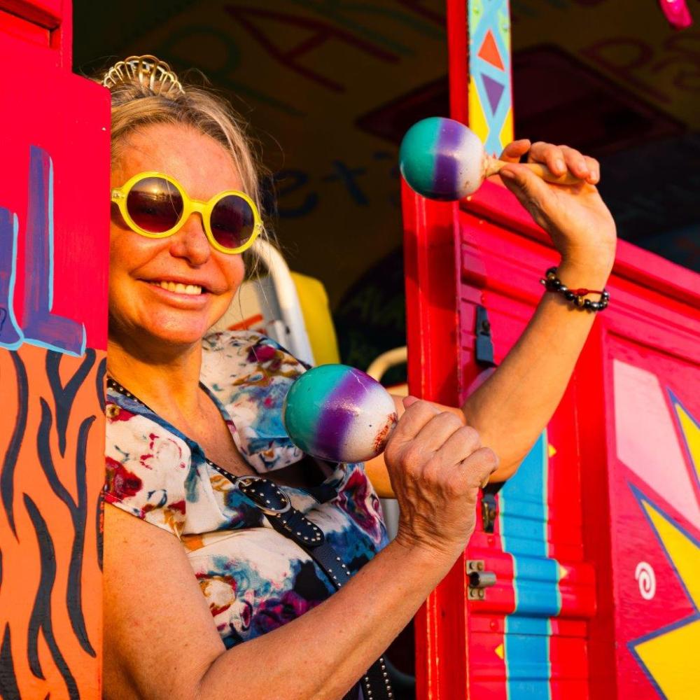 Woman with sunglasses and tiara holding maracas in a colorful setting.