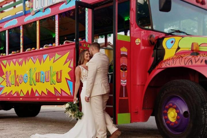 Bride and groom standing by a colorful party bus with 'Kukoo Kunuku' sign.