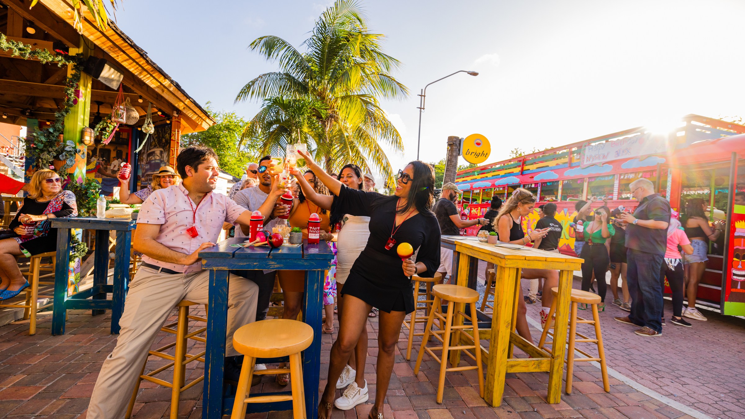 People enjoying drinks at colorful outdoor venue with a bright bus in the background.