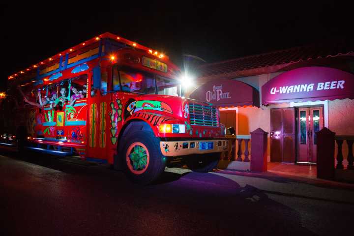 Colorful bus with people illuminated at night near bar with 'U-Wanna Beer' sign.