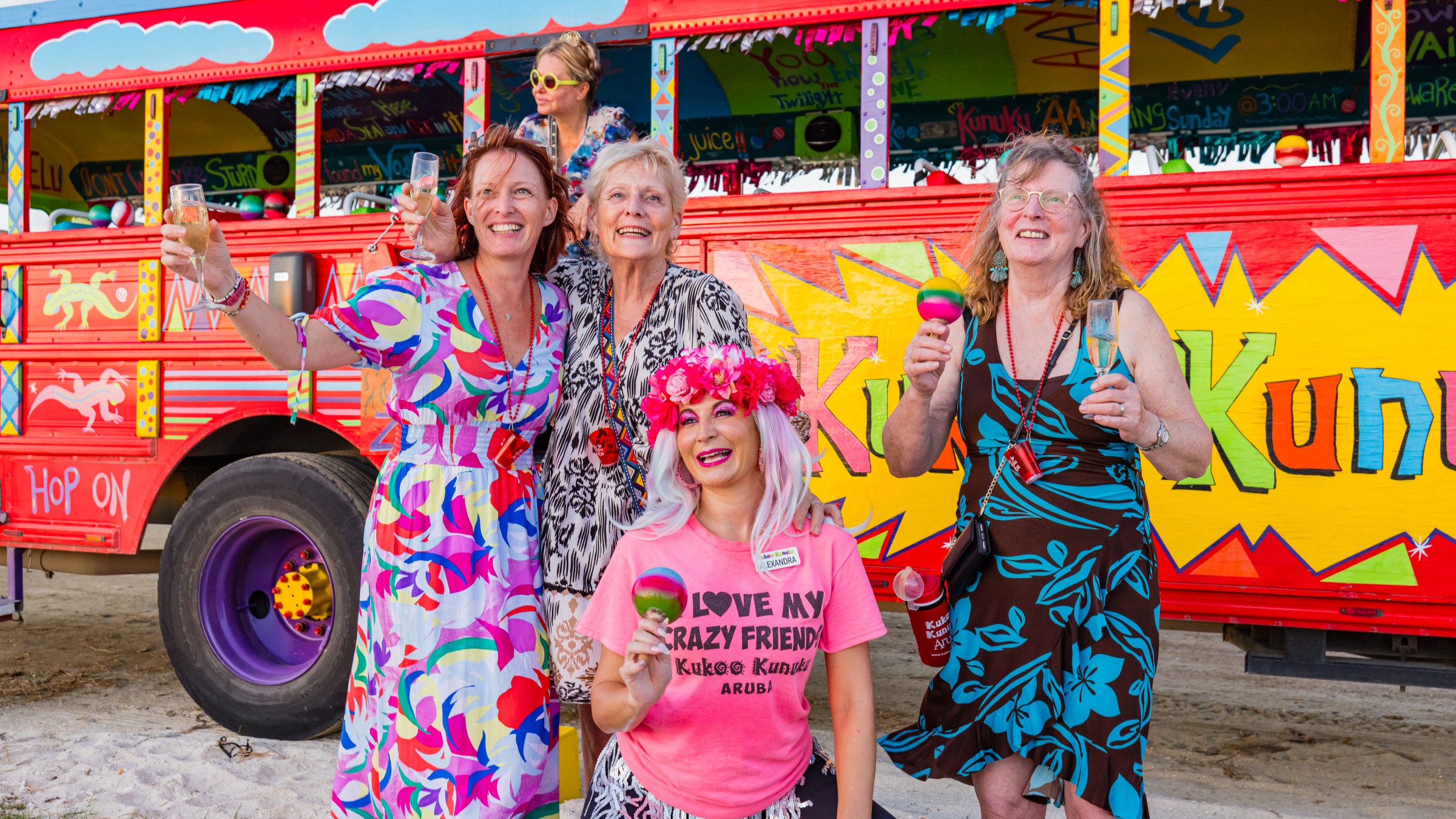 Four joyful women in colorful outfits pose in front of a vibrant, painted party bus.
