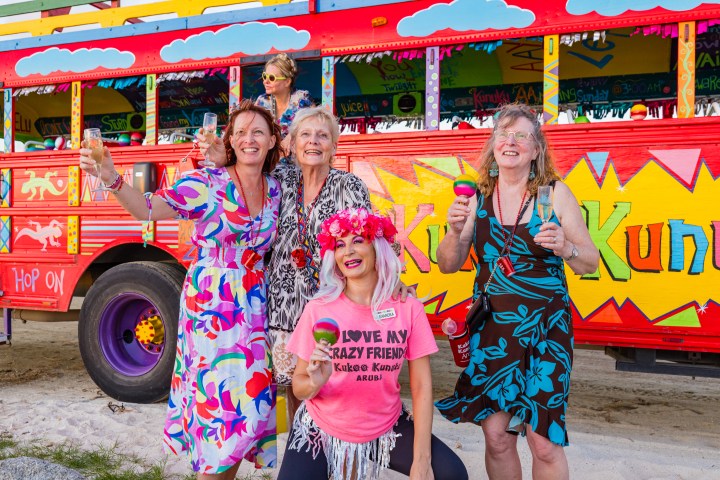 Four joyful women in colorful outfits pose in front of a vibrant, painted party bus.