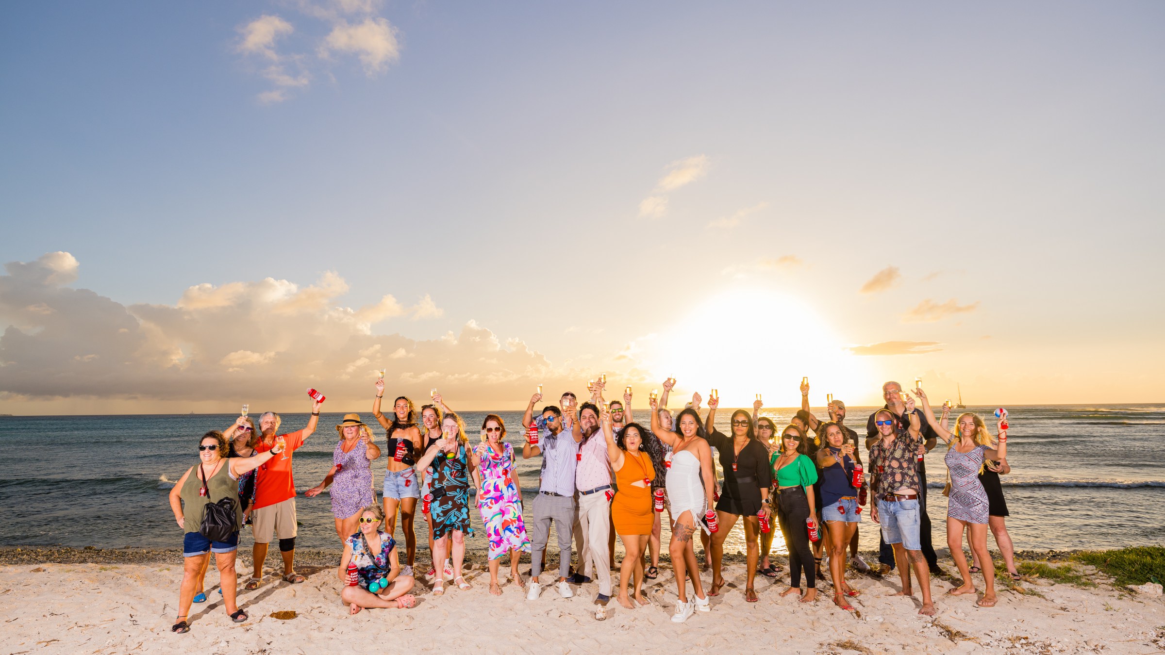 Group of people on a beach at sunset, holding drinks and smiling.