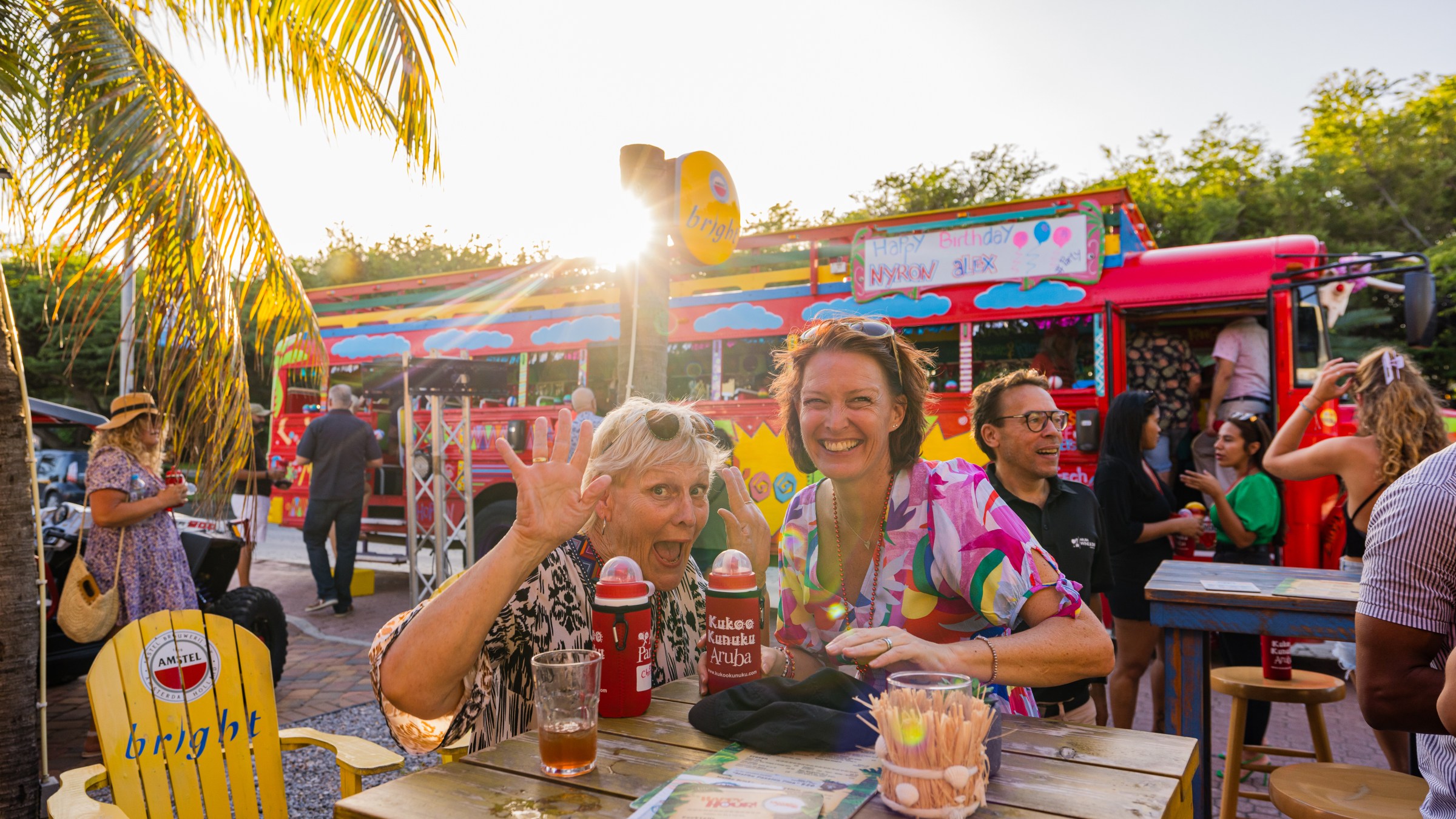 Two people smiling at an outdoor party with a colorful bus and palm trees in the background.