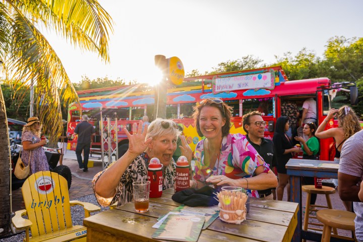 Two people smiling at an outdoor party with a colorful bus and palm trees in the background.