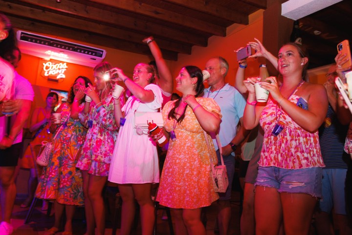 People in colorful clothes enjoying a lively event, holding drinks and phones, with neon signage in background.