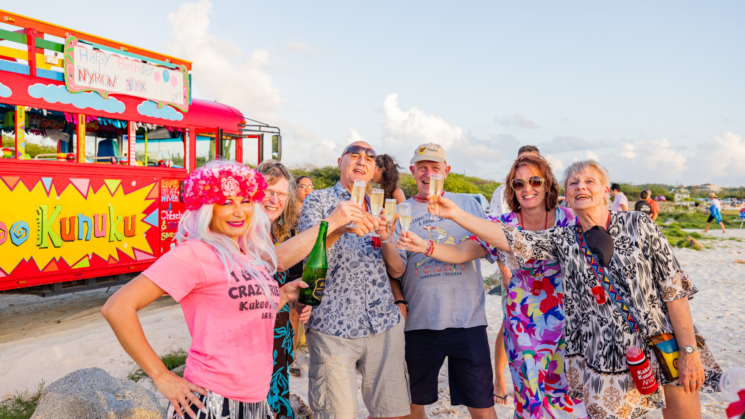 Group of people celebrating with drinks on a beach near a colorful bus.