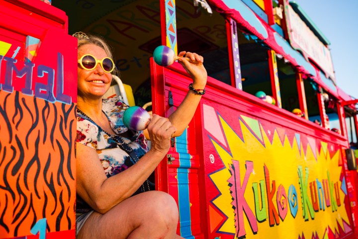 Woman in sunglasses holding maracas on a colorful bus with vibrant patterns.