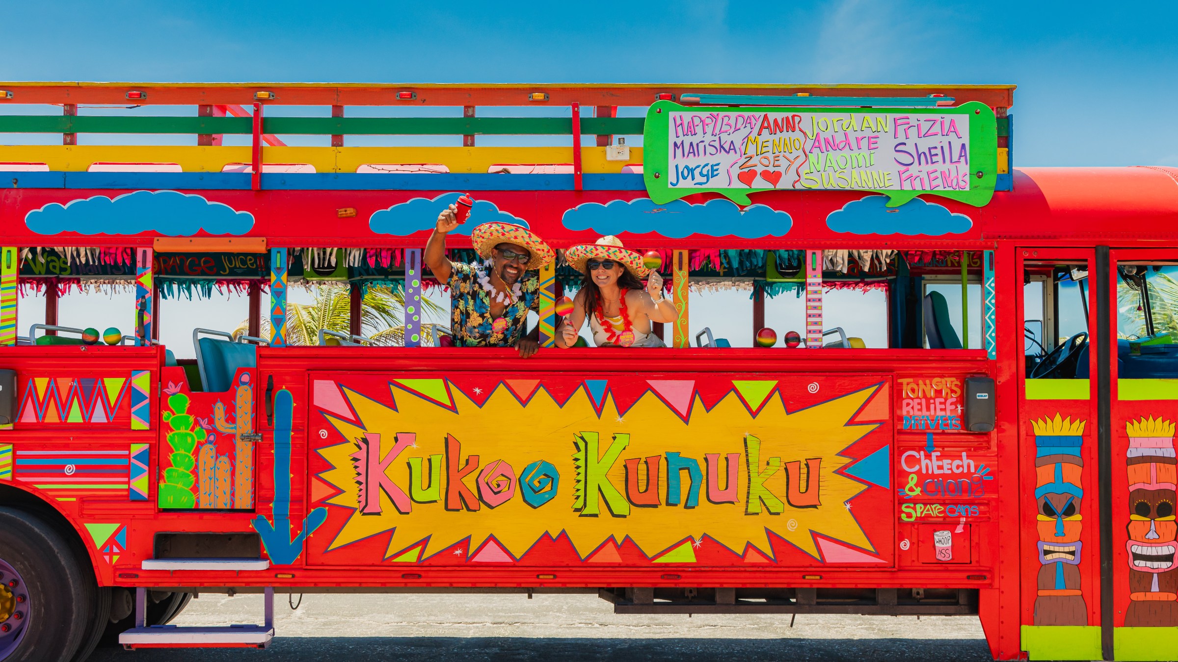 Colorful Kukoo Kunuku bus with two people wearing hats, waving through window, against a blue sky.