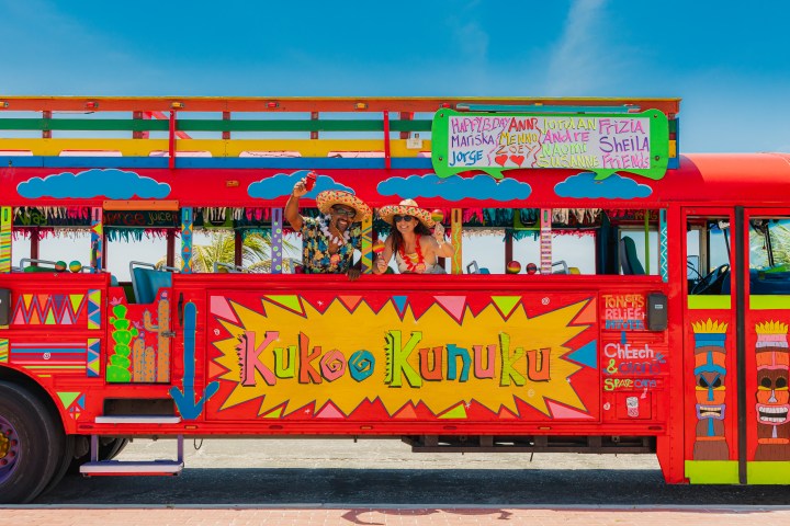 Colorful Kukoo Kunuku bus with two people wearing hats, waving through window, against a blue sky.