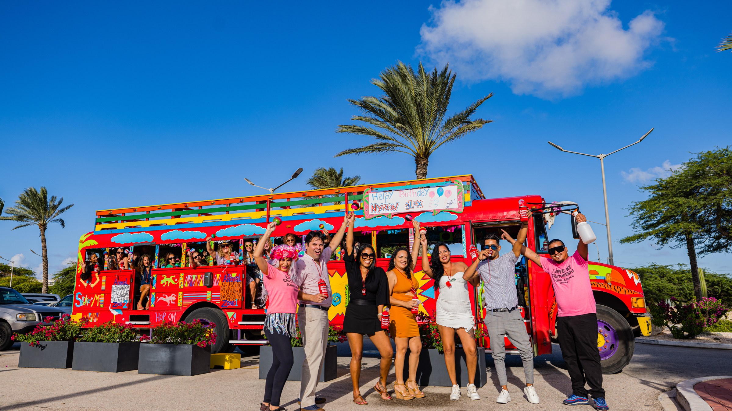 People celebrating in front of a colorful party bus with palm trees in the background.
