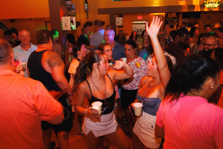 Crowded party scene with people dancing and holding drinks in a lively bar atmosphere.