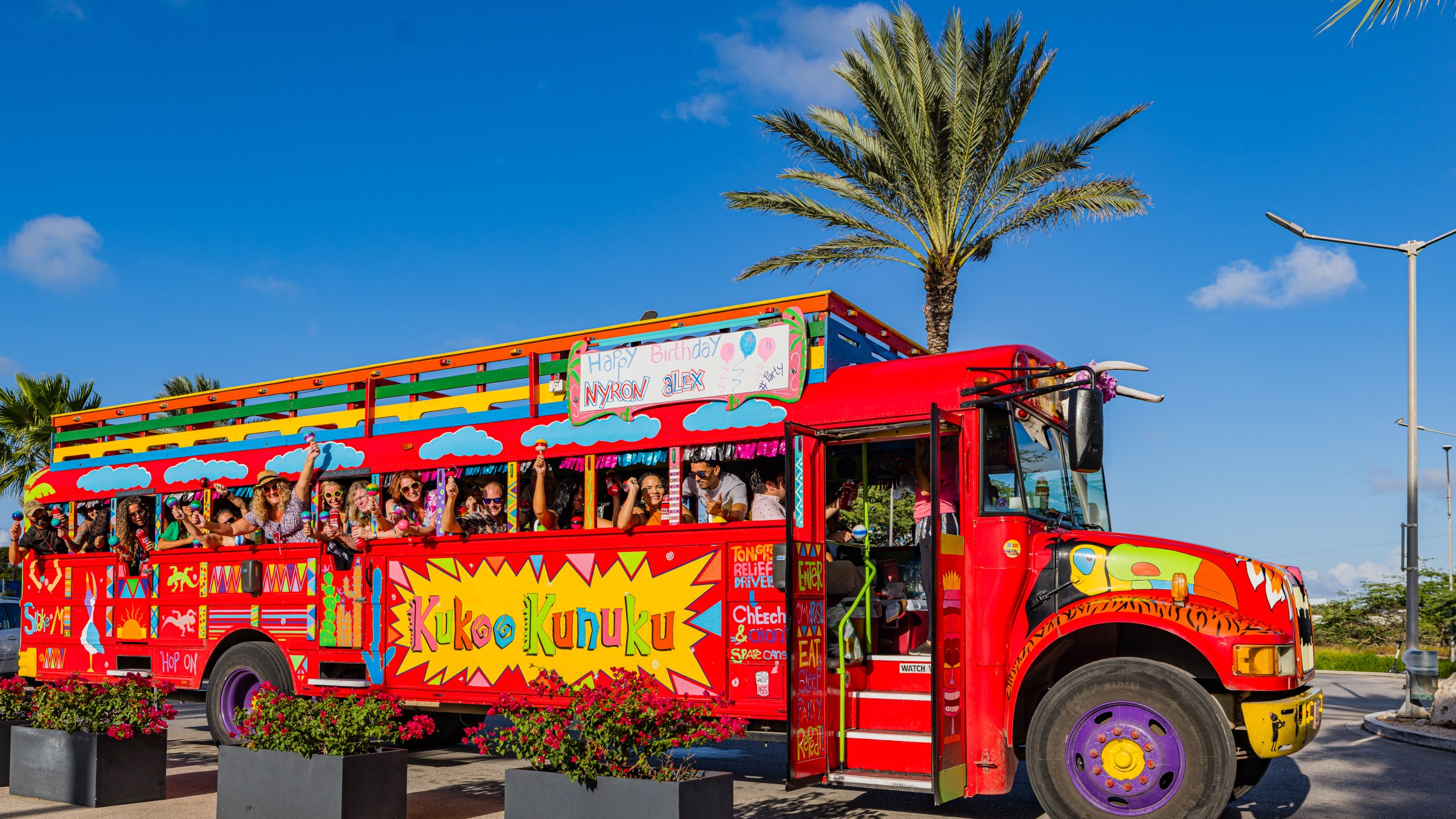 Colorful open-air party bus with palm trees and people onboard waving, under a clear blue sky.