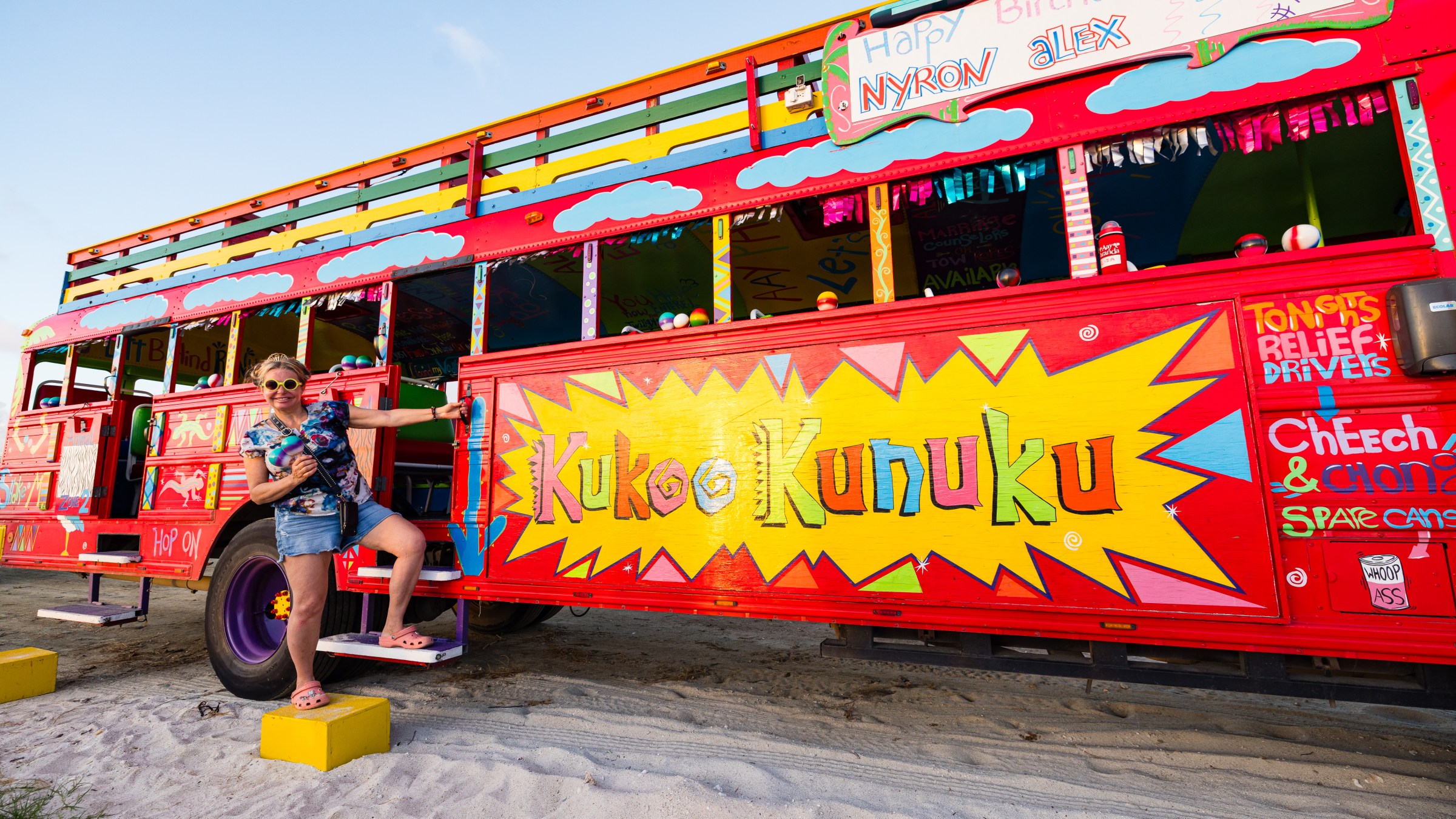 Woman in sunglasses posing with a colorful painted bus named 'Kukoo Kunuku' on a sandy beach.