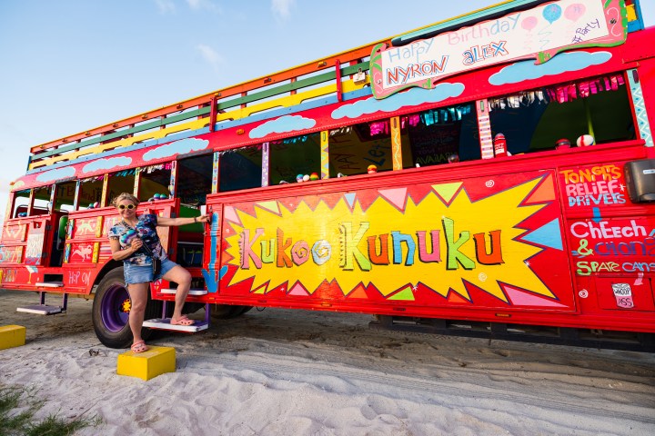 Woman in sunglasses posing with a colorful painted bus named 'Kukoo Kunuku' on a sandy beach.