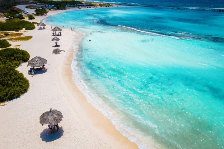 Aerial view of a tropical beach with turquoise water and palm thatched umbrellas on the sand.