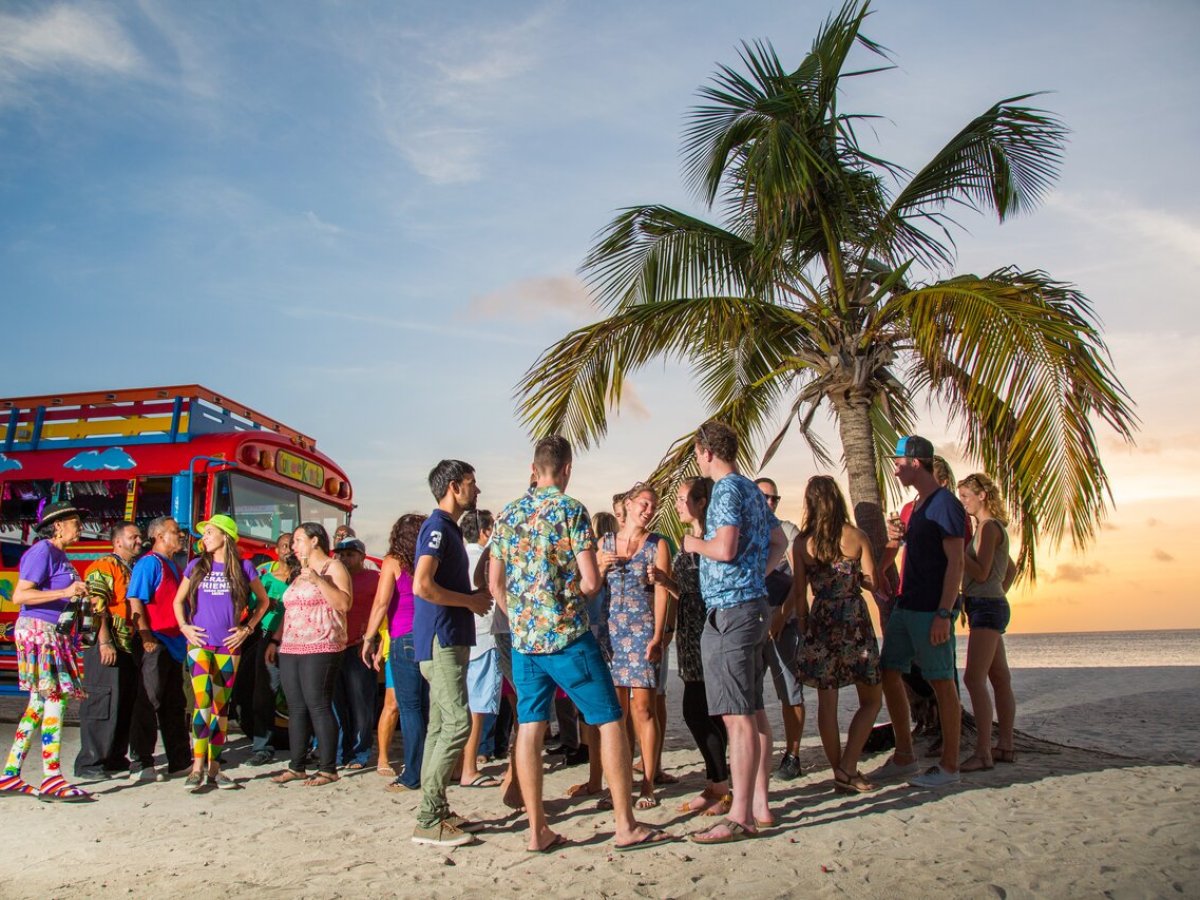 Group of people socializing on a beach near a colorful bus and a palm tree at sunset.