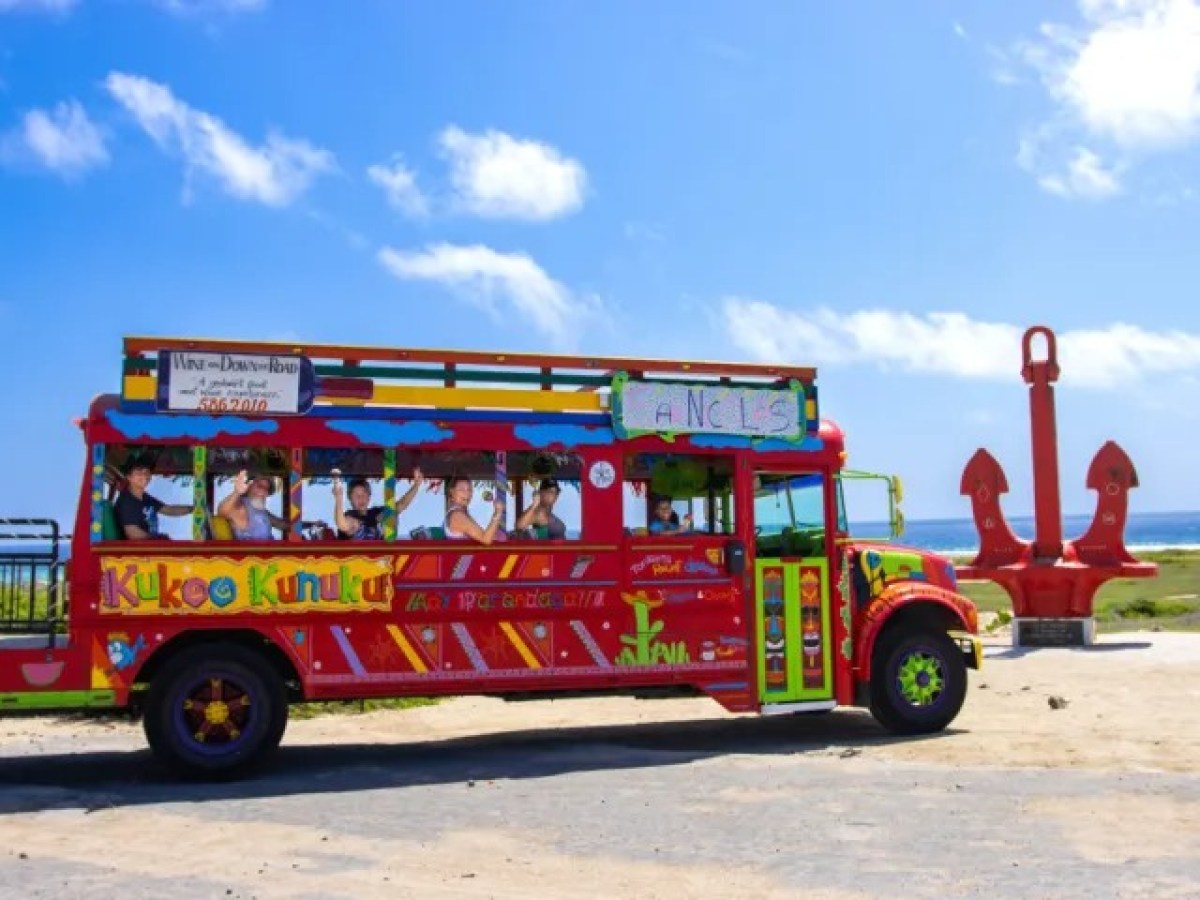 Colorful bus with passengers and large red anchors at beach under blue sky.