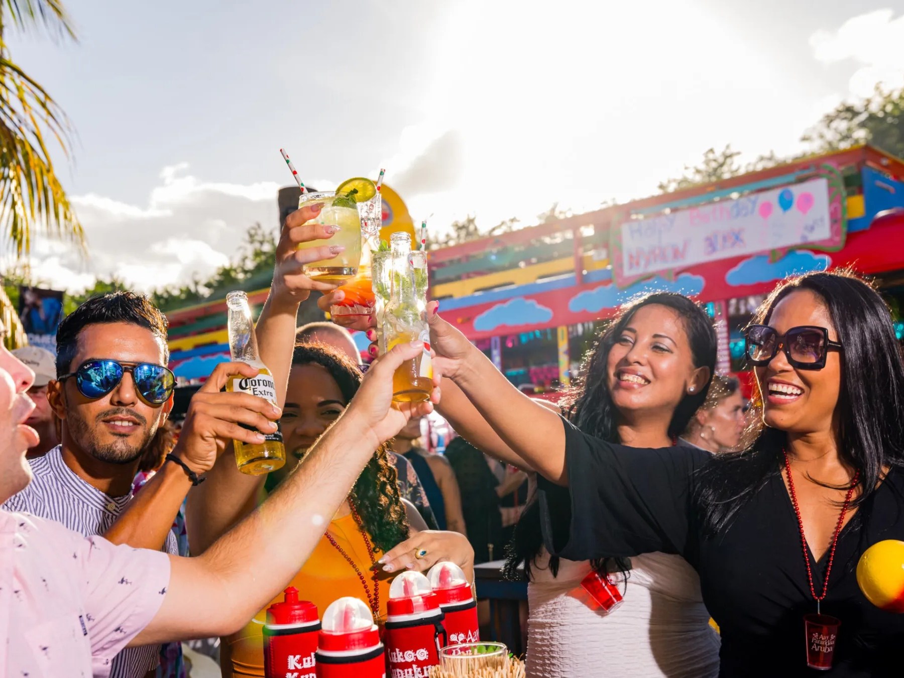 People at a lively outdoor party cheering with drinks, bright background, palm tree, and colorful setting.