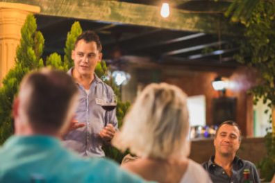 Person speaking to a group at an outdoor gathering, holding a wine glass.