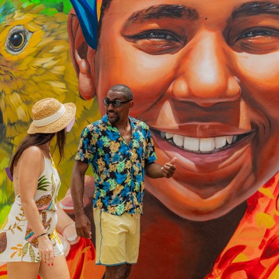 Two people talking in front of a colorful mural featuring a large face and a parrot.